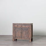 A rustic, weathered brown wooden side cabinet with two small drawers on top and two doors below, set against a plain light gray wall and gray floor.