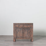 A rustic two-door wooden cabinet with two drawers on top, featuring a weathered brown finish and small round metal knobs, set against a plain white wall and gray floor.