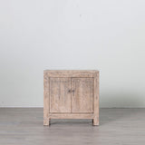 A small rustic light gray wooden cabinet with two doors and small metal knobs, set against a plain light gray wall and gray floor.