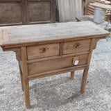 A rustic, light brown wooden writing desk or console table with two drawers and small metal knobs, photographed in what appears to be a workshop or furniture store.