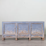 A rustic three-door sideboard cabinet with a distressed light blue and brown wood finish, set against a plain light gray wall and gray floor.