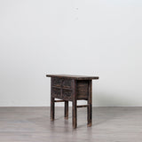 A small dark wooden side table with carved drawer fronts and four legs, photographed in a minimalist studio setting with a light gray wall and gray floor.
