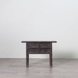 A dark, weathered wooden side table with four drawers and carved drawer fronts, set against a plain white wall and light gray floor.