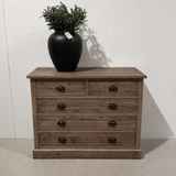 Wooden dresser with black drawers and a black vase with greenery on top against a white wall.