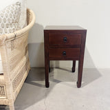 Wooden nightstand with two drawers next to a wicker chair against a white wall.