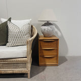 Wooden side table with a textured lamp next to a wicker chair with patterned cushions.