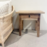 Wooden side table with a drawer next to a wicker chair against a white wall.