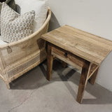 Wooden side table next to a wicker chair with patterned cushions on a concrete floor.