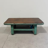 Wooden table with green base on a concrete floor against a white wall