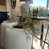 White cylindrical planters with dried plants on a wooden surface, with a lamp and wall decor in the background.