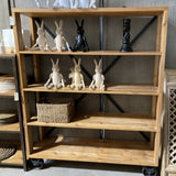 A rustic wooden 4-tier bookcase with black metal X-brace supports and caster wheels, styled with rabbit figurines and a wicker basket in a furniture showroom.