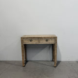 A rustic two-drawer console table with a distressed, weathered finish, set against a plain white wall and concrete floor.