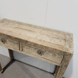 A rustic, distressed wooden console table with two drawers and metal knobs, photographed in a warehouse setting against a white wall.