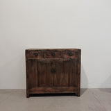 A rustic dark brown wooden sideboard cabinet with three drawers on top and two doors below, photographed against a plain off-white wall and concrete floor.