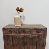 A rustic dark brown wooden sideboard cabinet with three drawers and two lower doors, styled with a beige and white ceramic vase containing dried brown flowers against a plain white wall.