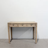 A rustic three-drawer console table made of light-colored weathered wood, featuring small metal drawer pulls and four tapered legs, set against a plain white wall and concrete floor.