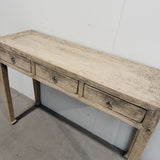 A rustic three-drawer console table made of weathered light wood with dark metal drawer pulls, photographed against a plain white wall and concrete floor.