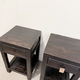 Two dark brown wooden end tables with distressed finishes, featuring a single drawer and a lower shelf, photographed against a plain off-white wall.