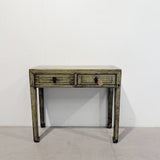 A distressed vintage-style writing desk with two drawers, featuring a weathered green and brown painted finish with dark metal drawer pulls, set against a plain white wall and concrete floor.