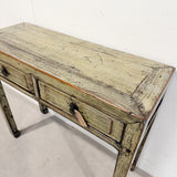 A rustic, distressed wooden console table with a weathered green and brown finish, featuring two drawers with dark metal knobs and a hanging price tag, photographed against a plain white wall.