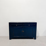 A dark blue wooden sideboard cabinet with two drawers on top and two doors below, featuring small round metal knobs, set against a plain white wall and light concrete floor.