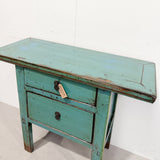 A distressed turquoise green wooden nightstand with two drawers, a single round knob, and a keyhole detail on the front, set against a plain white wall.
