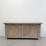 An antique carved wooden chest or sideboard with intricate geometric patterns, set against a plain white wall and concrete floor.