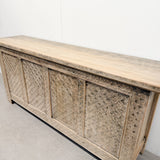 A long carved wooden sideboard or credenza with a light natural wood top and four textured carved front panels, photographed indoors against a plain white wall and concrete floor.