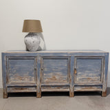 A rustic three-door sideboard or credenza in distressed blue wood with a weathered brown and beige finish, styled with a ceramic lamp on top against a plain off-white wall.