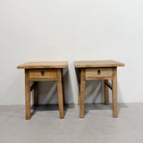 Two rustic wooden end tables with single drawers and black metal ring pulls, set against a plain white wall and concrete floor.