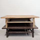 A rustic wooden coffee table with a light natural wood top, a dark brown lower shelf, and a middle open storage shelf, set against a plain white wall.