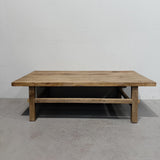 A rustic rectangular wooden coffee table with a thick natural wood top and a lower support rail, set against a plain white wall and gray concrete floor.