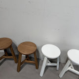 Three small stools lined up against a plain light gray wall and concrete floor, with two wooden stools on the left and one white stool on the right.