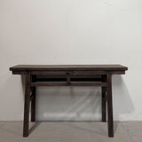 A dark brown wooden console table with a rectangular top, two drawers, and four legs, set against a plain off-white wall and concrete floor.