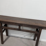 A dark brown wooden console table with a long rectangular top and open lower shelf, photographed against a plain white wall and light concrete floor.