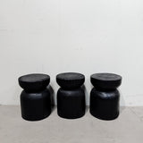 Three black wooden barrel-shaped stools with round cut-stump style tops, arranged side by side against a white wall and light concrete floor.