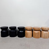 Six rustic wooden stools arranged in two groups of three, featuring a mix of black-painted and natural light wood finishes against a plain off-white wall and concrete floor.