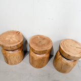 Three rustic wooden log stools with thick circular tree-stump tops and cylindrical bases, arranged against a plain light gray wall and concrete floor.