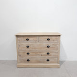 A rustic light oak wood five-drawer chest with two smaller top drawers and five larger drawers below, featuring black metal knobs, set against a plain white wall and concrete floor.