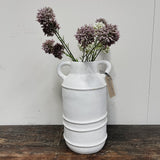 A white terracotta vase with two handles, containing pink and white flowers, placed on a wooden surface against a plain background.
