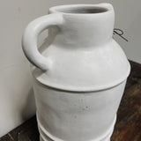 A white terracotta vase with two handles, containing pink and white flowers, placed on a wooden surface against a plain background.