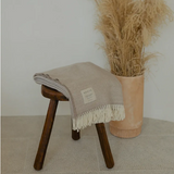 Beige blanket with white fringe draped over a wooden stool next to a vase with dried plants on a light-colored floor.