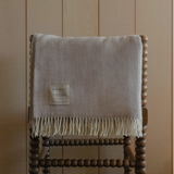 Gray throw blanket with fringes on a wooden chair against a beige wall.