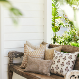 A rustic wooden bench on a porch is styled with several decorative throw pillows in neutral beige, brown, and cream tones, set against a white shiplap wall with green foliage in the background.