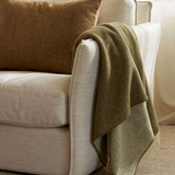 A textured olive green throw blanket with fringe is draped over the arm of a cream-colored upholstered armchair, next to a brown woven pillow.