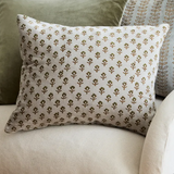 A square throw pillow with a repeating brown and olive green floral block print on a cream fabric, styled on a beige armchair with green and patterned cushions behind it.
