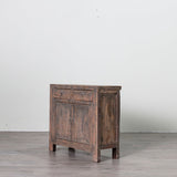 A rustic dark brown wooden side cabinet with two small drawers on top and two larger doors below, shown on a light gray floor against a white wall.