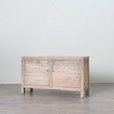 A rustic, distressed light gray wooden sideboard cabinet with two doors and small legs, set against a plain light gray wall and gray floor.