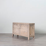 A distressed light gray wood sideboard cabinet with two doors and small legs, shown in a minimalist studio setting with a gray floor and white wall.