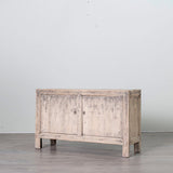 A rustic, distressed light gray wood sideboard cabinet with two doors and small legs, shown in a minimalist studio setting with a gray floor and white wall.